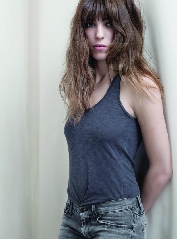 Eine Frau mit langen, gewellten Haaren und einem grauen Tanktop lehnt an einer Wand.