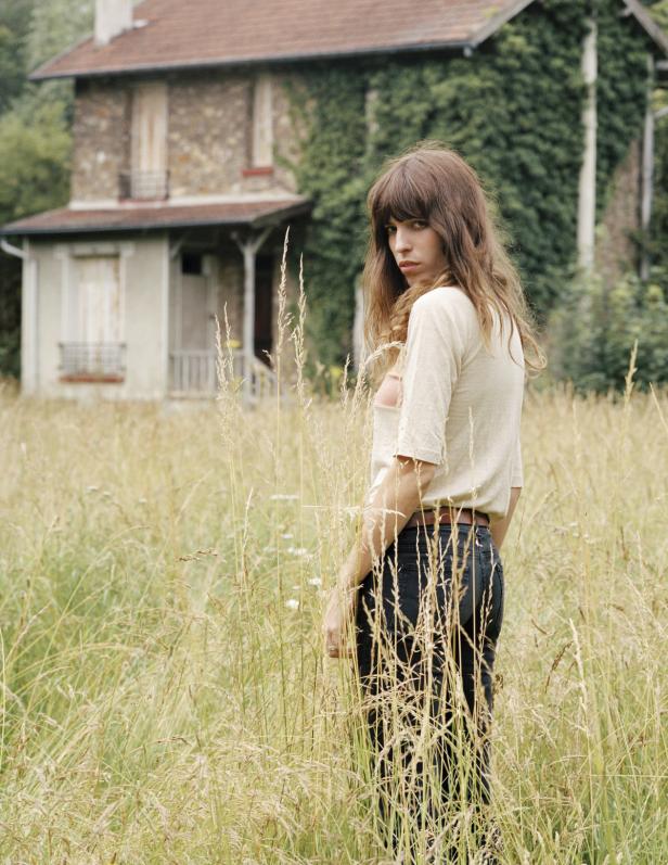 Eine Frau steht in einem hohen Grasfeld vor einem alten Haus.