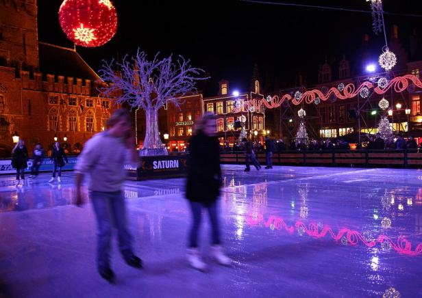 Menschen laufen auf einer Eisbahn vor weihnachtlich beleuchteten Gebäuden.