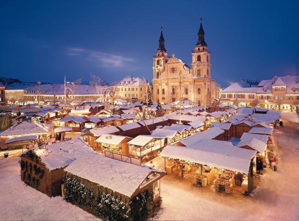 Der Weihnachtsmarkt in Ravensburg mit schneebedeckten Buden und der Stadtpfarrkirche im Hintergrund.