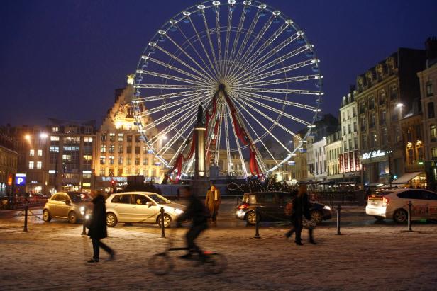 Ein belebter Platz in Lille bei Nacht mit einem großen Riesenrad im Hintergrund.