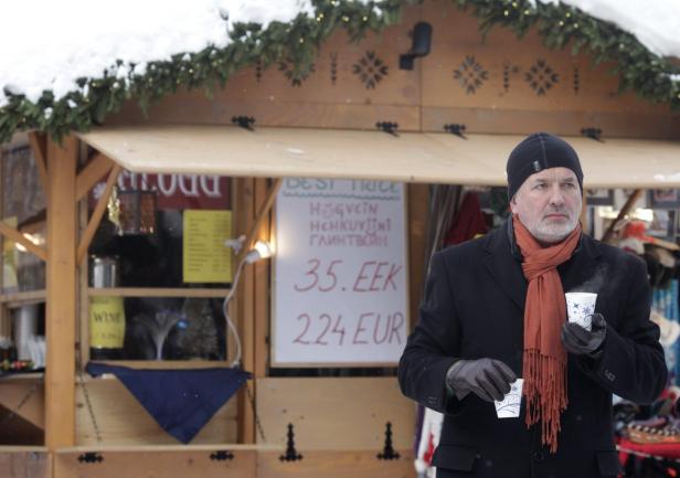 Ein Mann mit Mütze und Schal hält einen Becher vor einem Verkaufsstand auf einem Weihnachtsmarkt.