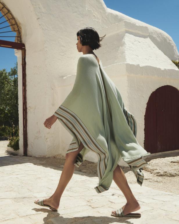 Eine Frau geht in einem hellgrünen Poncho mit Streifen vor einem weißen Gebäude entlang.