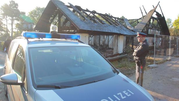 Ein Polizist steht vor einem ausgebrannten Gebäude, im Vordergrund ein Polizeiauto.
