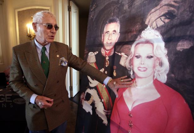 Ein Mann in einem karierten Sakko posiert vor einem Porträt von Zsa Zsa Gabor und ihrem Ehemann Prinz Frédéric von Anhalt.