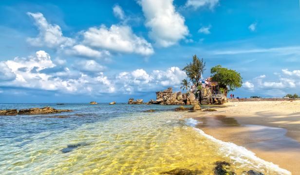 Ein Strand mit einem kleinen Tempel auf einem Felsen unter blauem Himmel mit Wolken.