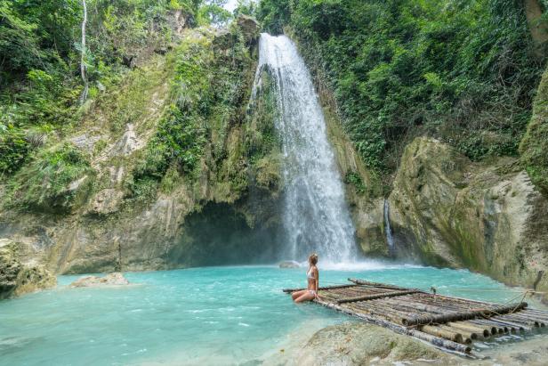 Eine Frau sitzt auf einem Bambusfloß vor einem Wasserfall mit türkisfarbenem Wasser.