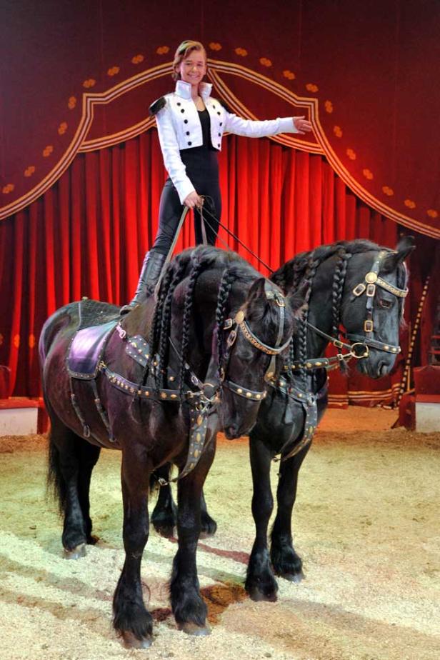Eine junge Frau steht auf zwei geschmückten, schwarzen Pferden in einem Zirkus.