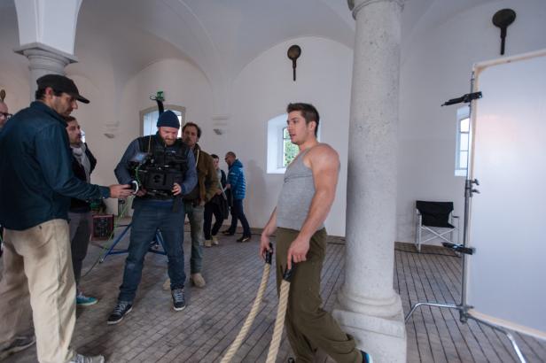 Ein Mann im grauen Tanktop zieht an Seilen, während ein Filmteam ihn filmt.