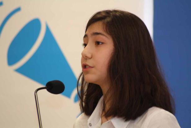 Ein junges Mädchen spricht in ein Mikrofon vor einem blauen Hintergrund.