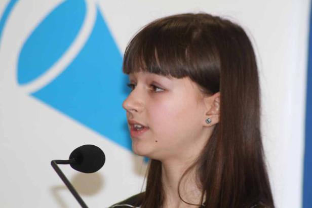 Ein junges Mädchen spricht in ein Mikrofon vor einem blauen und weißen Hintergrund.