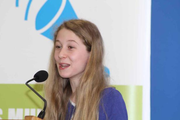 Eine junge Frau spricht an einem Mikrofon vor einem farbigen Hintergrund.