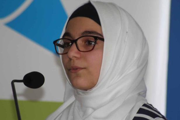 Eine junge Frau mit Brille und Hijab spricht an einem Mikrofon.