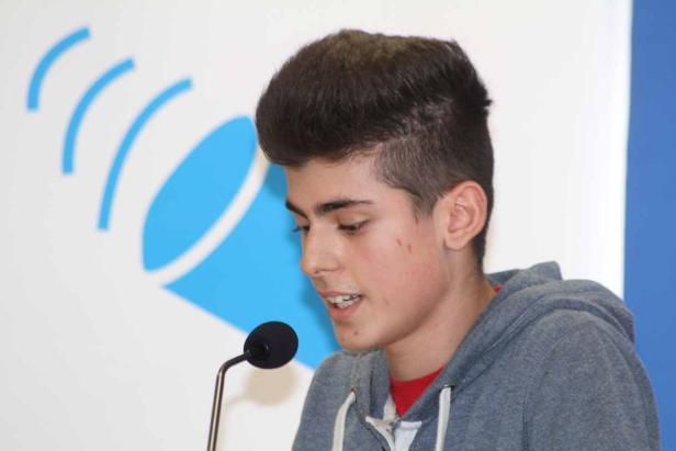 Ein junger Mann spricht in ein Mikrofon vor einem blauen Logo.