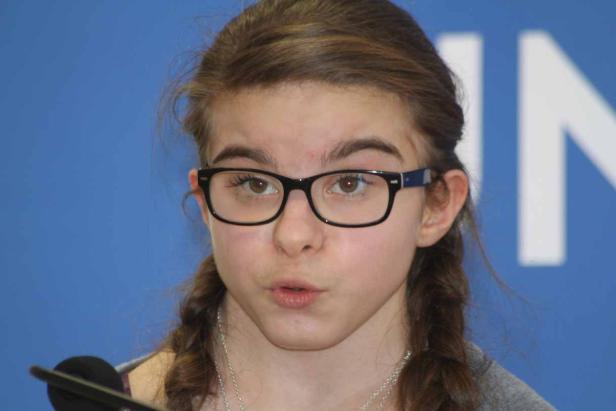 Ein junges Mädchen mit Brille und Zöpfen spricht in ein Mikrofon.