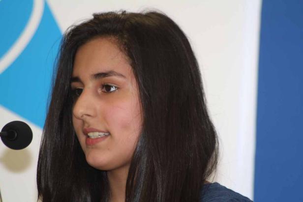 Eine junge Frau mit dunklen Haaren spricht vor einem blauen Hintergrund.