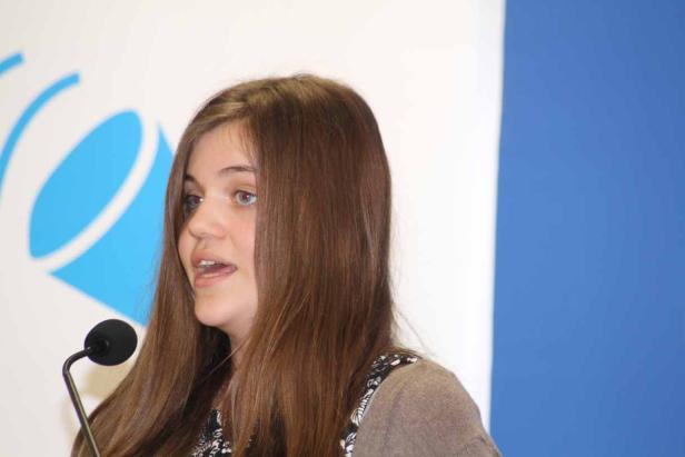 Eine junge Frau spricht am Mikrofon vor einem blauen und weißen Hintergrund.