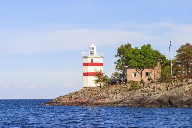 Ein rot-weiß gestreifter Leuchtturm auf einer felsigen Insel mit einem Haus und der schwedischen Flagge.