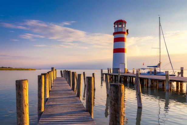 Ein rot-weiß gestreifter Leuchtturm steht an einem See neben einem Steg und einem Segelboot.