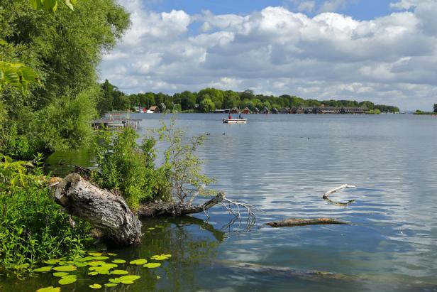 Ein ruhiger See mit einem kleinen Boot und Häusern am Ufer unter einem bewölkten Himmel.