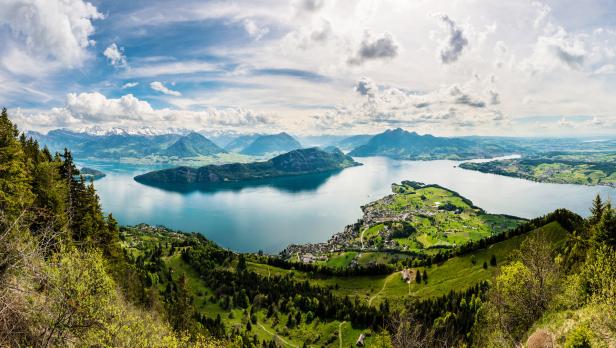 Panoramablick auf einen See und grüne Hügel mit schneebedeckten Bergen im Hintergrund.