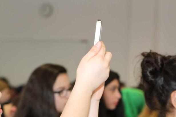 Eine Hand hält ein Smartphone in die Höhe, im Hintergrund sind verschwommene Gesichter zu sehen.