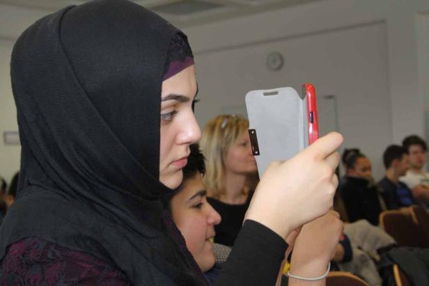 Eine Frau mit Hijab benutzt ihr Smartphone in einem Raum voller Menschen.
