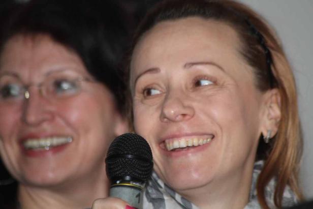 Eine Frau lächelt und hält ein Mikrofon in der Hand, während eine andere Frau im Hintergrund steht.