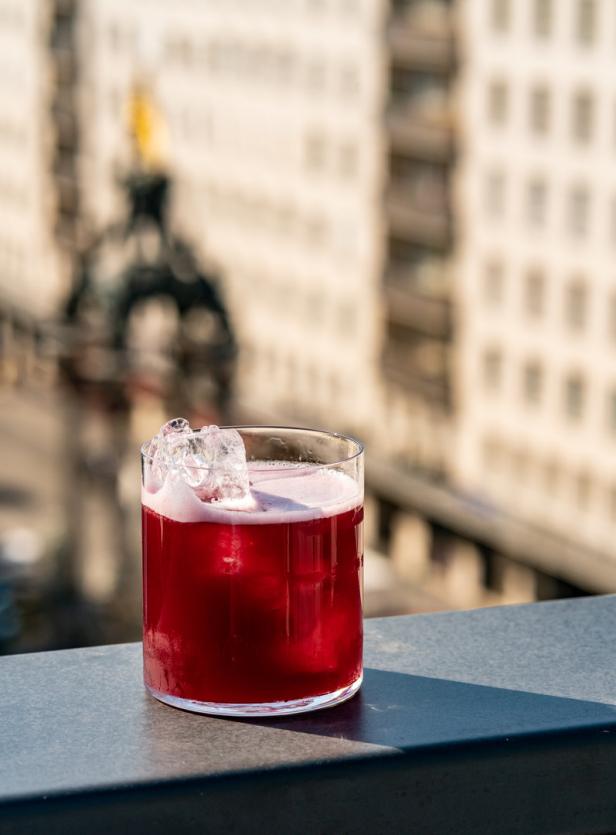 Ein roter Cocktail mit Eiswürfeln auf einer Dachterrasse mit Blick auf die Stadt.
