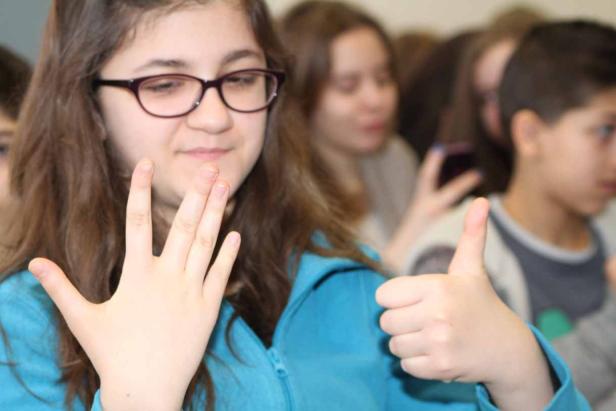 Ein Mädchen mit Brille zeigt mit einer Hand die Zahl fünf und mit der anderen einen Daumen nach oben.