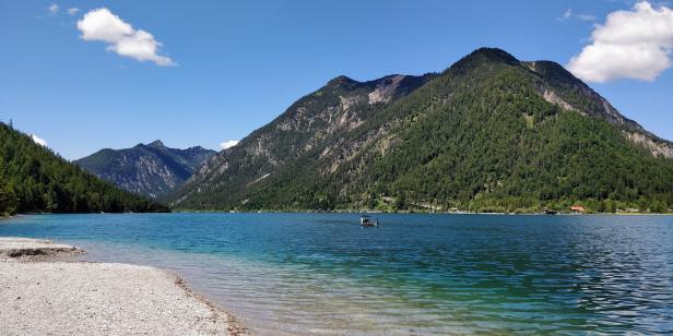 Ein ruhiger See umgeben von bewaldeten Bergen unter blauem Himmel.