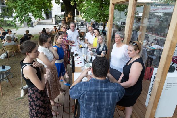 Eine Gruppe von Menschen steht um einen Tisch im Freien, möglicherweise bei einer Weinprobe.