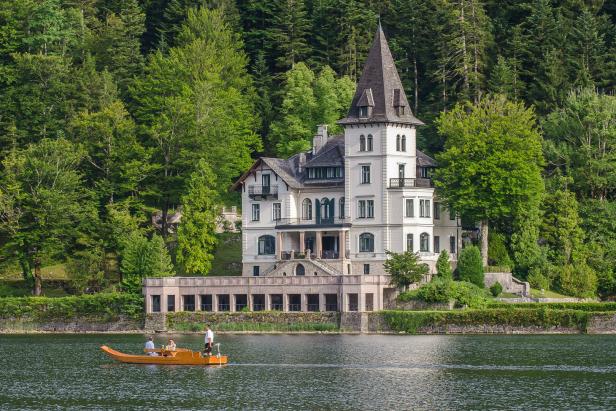 Ein Ruderboot fährt auf einem See vor einem großen, weißen Haus mit Turm.