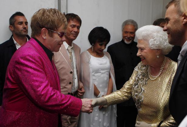 Königin Elisabeth II. schüttelt Elton John die Hand, umgeben von weiteren Personen.