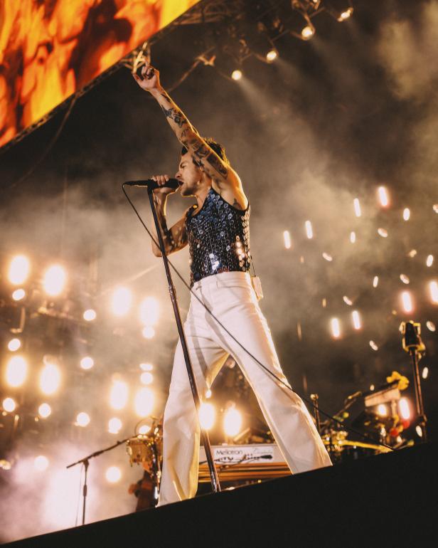 Harry Styles tritt in einem glitzernden Oberteil und einer weißen Hose auf einer Bühne auf.