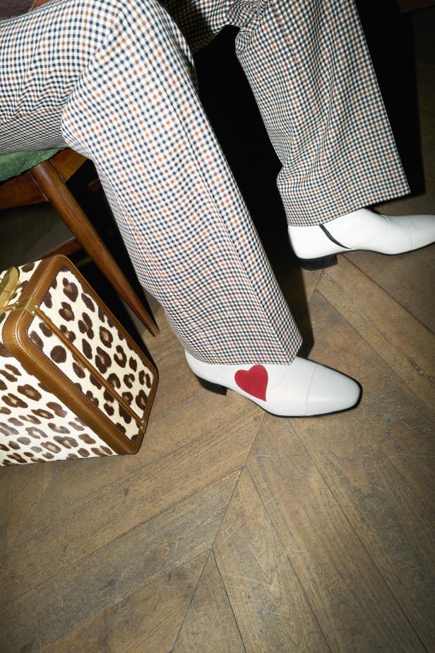 Eine Person mit karierter Hose und weißen Schuhen mit rotem Herz sitzt auf einem Stuhl neben einer Handtasche mit Leopardenmuster.