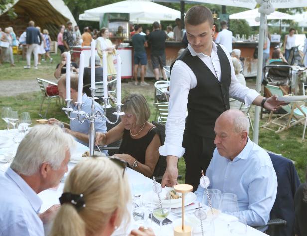 Ein Kellner serviert Speisen an einem Tisch im Freien, an dem mehrere Personen sitzen.