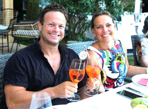 Ein lächelndes Paar sitzt an einem Tisch und hält Aperol Spritz Gläser in der Hand.