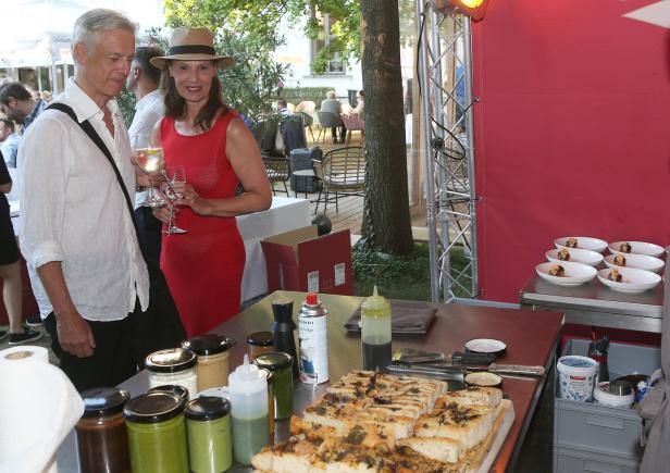 Eine Frau mit Hut und ein Mann mit Getränken stehen an einem Tisch mit Speisen bei einer Veranstaltung.