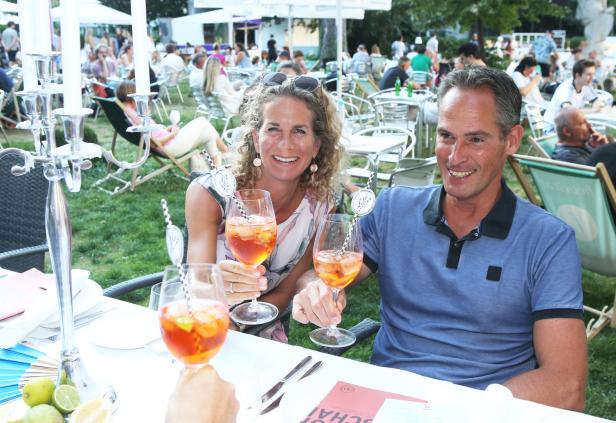 Ein Paar prostet mit Aperol Spritz in einem Garten, im Hintergrund weitere Gäste.