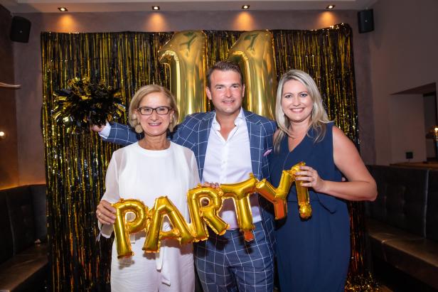 Drei Personen posieren mit goldenen „Party“-Ballons vor einem goldenen Lametta-Hintergrund.