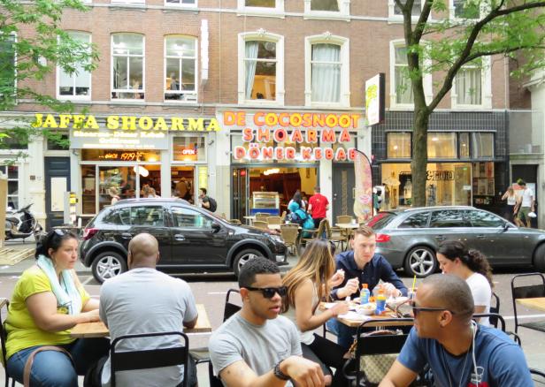 Menschen sitzen vor Restaurants in einer belebten Straße und essen.