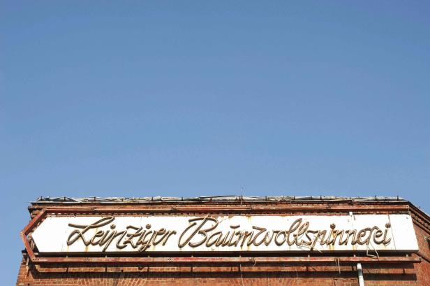 Das Schild „Leipziger Baumwollspinnerei“ an einem Backsteingebäude vor blauem Himmel.