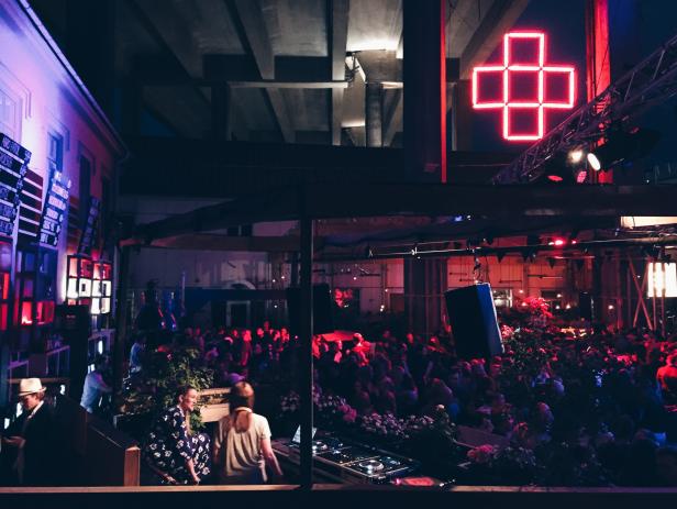 Eine Menschenmenge versammelt sich in einem beleuchteten Veranstaltungsort mit einer DJ-Konsole im Vordergrund.
