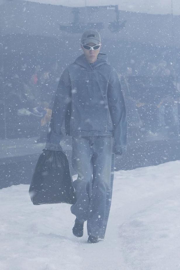 Eine Person in Jeanskleidung und Sonnenbrille trägt einen Müllsack durch den Schnee.