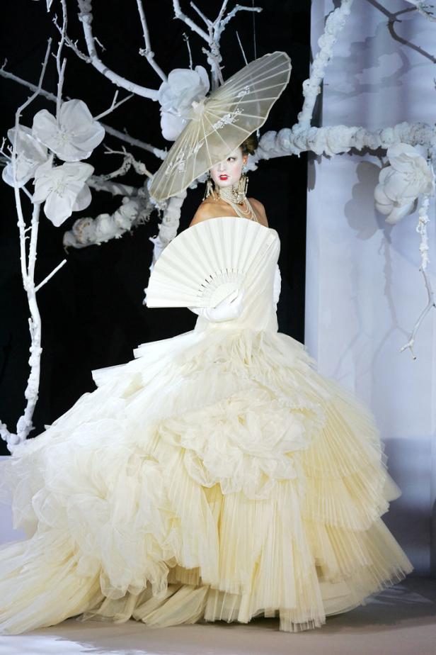 Eine Frau in einem weißen Ballkleid mit Fächer und einem Schirmchen im Haar vor einem floralen Hintergrund.