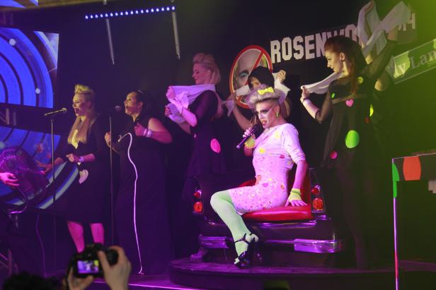 Eine Dragqueen sitzt auf einem Autositz während einer Bühnenperformance mit anderen Künstlerinnen.