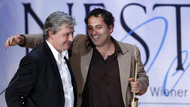 Zwei Männer in Anzügen umarmen sich, einer hält eine goldene Trophäe.