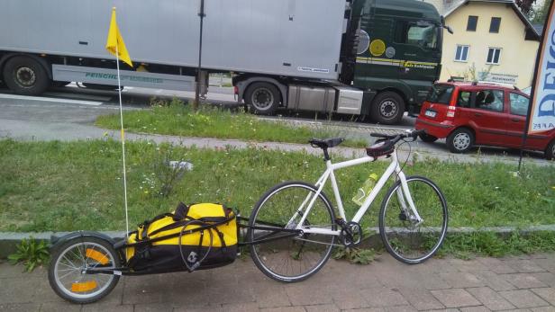 Ein weißes Fahrrad mit Anhänger und gelber Plane steht am Straßenrand.