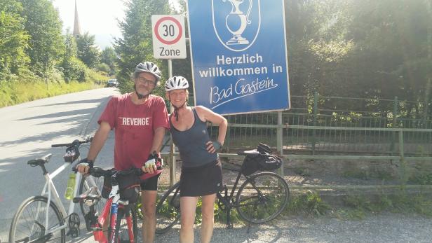Ein Paar mit Fahrrädern posiert vor einem „Willkommen in Bad Gastein“-Schild.